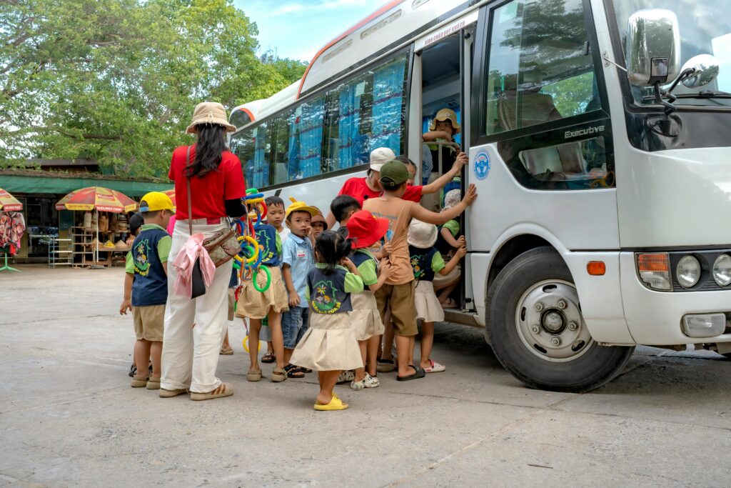 Group of children and adults boarding a bus, preparing for a school trip in a sunny outdoor setting.